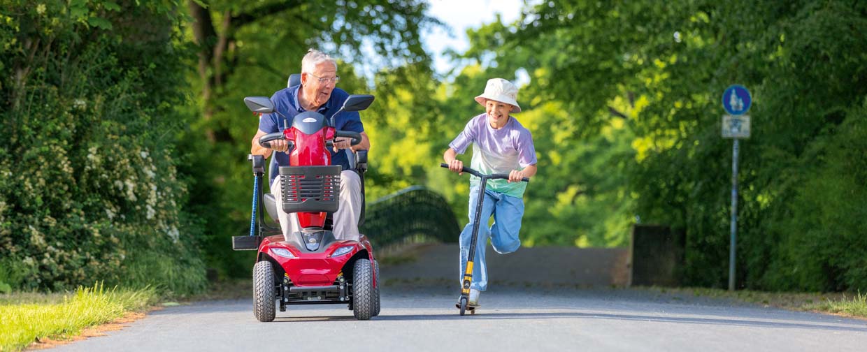 Fotografie zeigt &auml;lteren Mann auf rotem Elektromobil und Kind auf Tretroller, die nebeneinander auf einem von B&auml;umen ges&auml;umten Weg ein Wettrennen fahren.