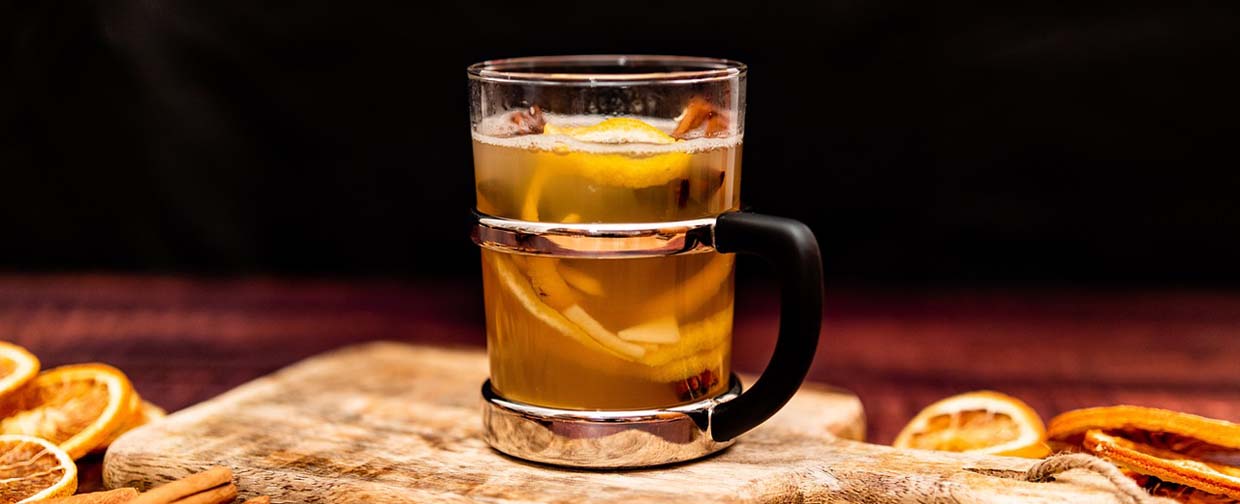 Ein Glasbecher mit alkoholfreiem Punsch steht auf einem Holzbrettchen. Im Punsch sind Zitronenschale und Ingwerstückchen sowie ein paar Gewürzblätter zu sehen.
