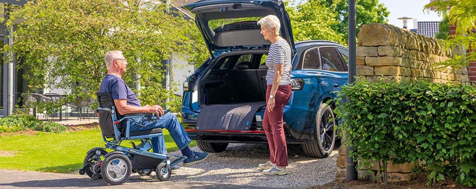 Die Fotografie zeigt einen Mann im Elektrorollstuhl und eine Frau vor dem geöffneten Kofferraum eines blauen Autos in einer Gartenumgebung. Die Szene vermittelt eine alltägliche Situation und rückt dabei Mobilitätshilfe sowie die Bedeutung eines barrierefreien Transports in den Mittelpunkt.
